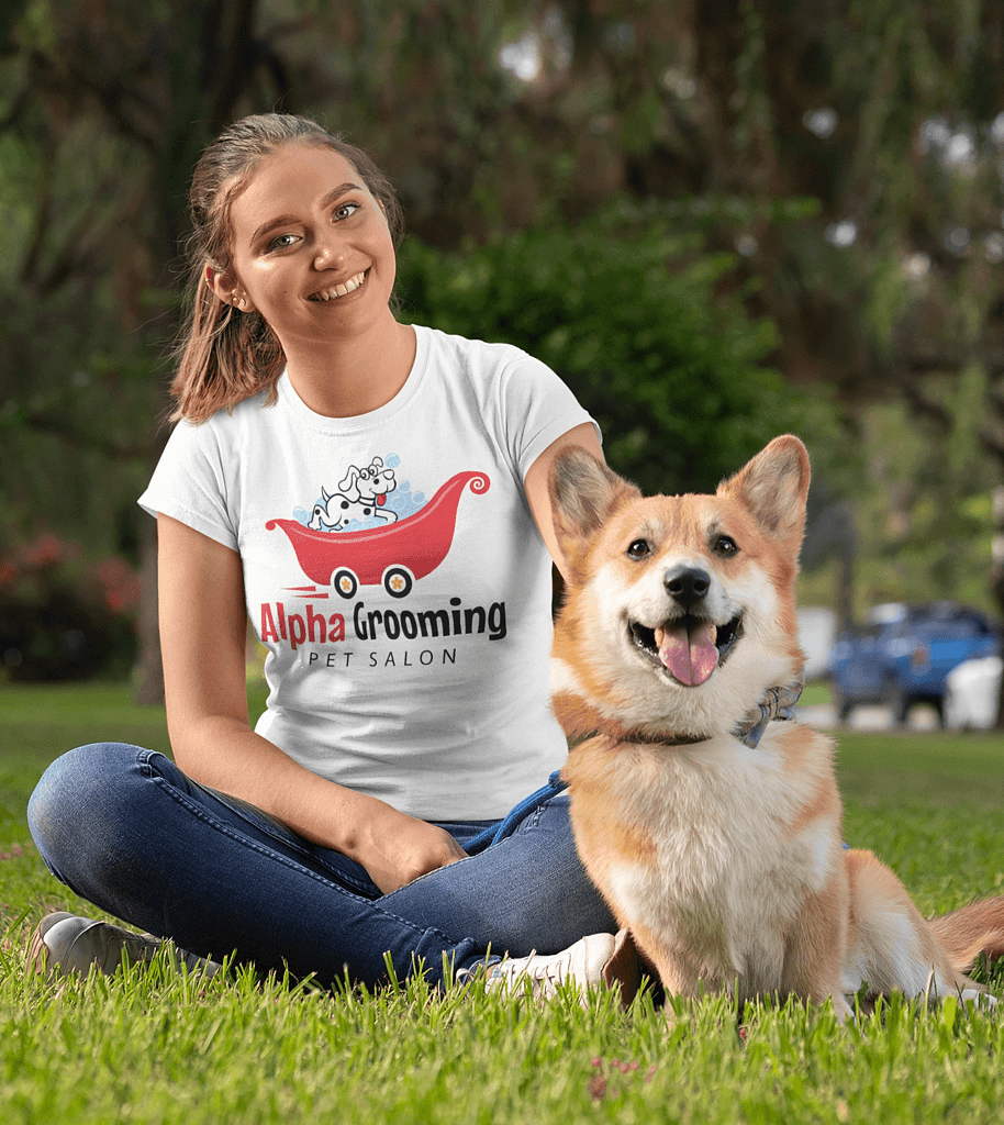 Woman sitting on grass with a smiling dog, wearing a white "Alpha Grooming Pet Salon" t-shirt; park setting with trees in the background—perfectly capturing the joy of mobile pet grooming.